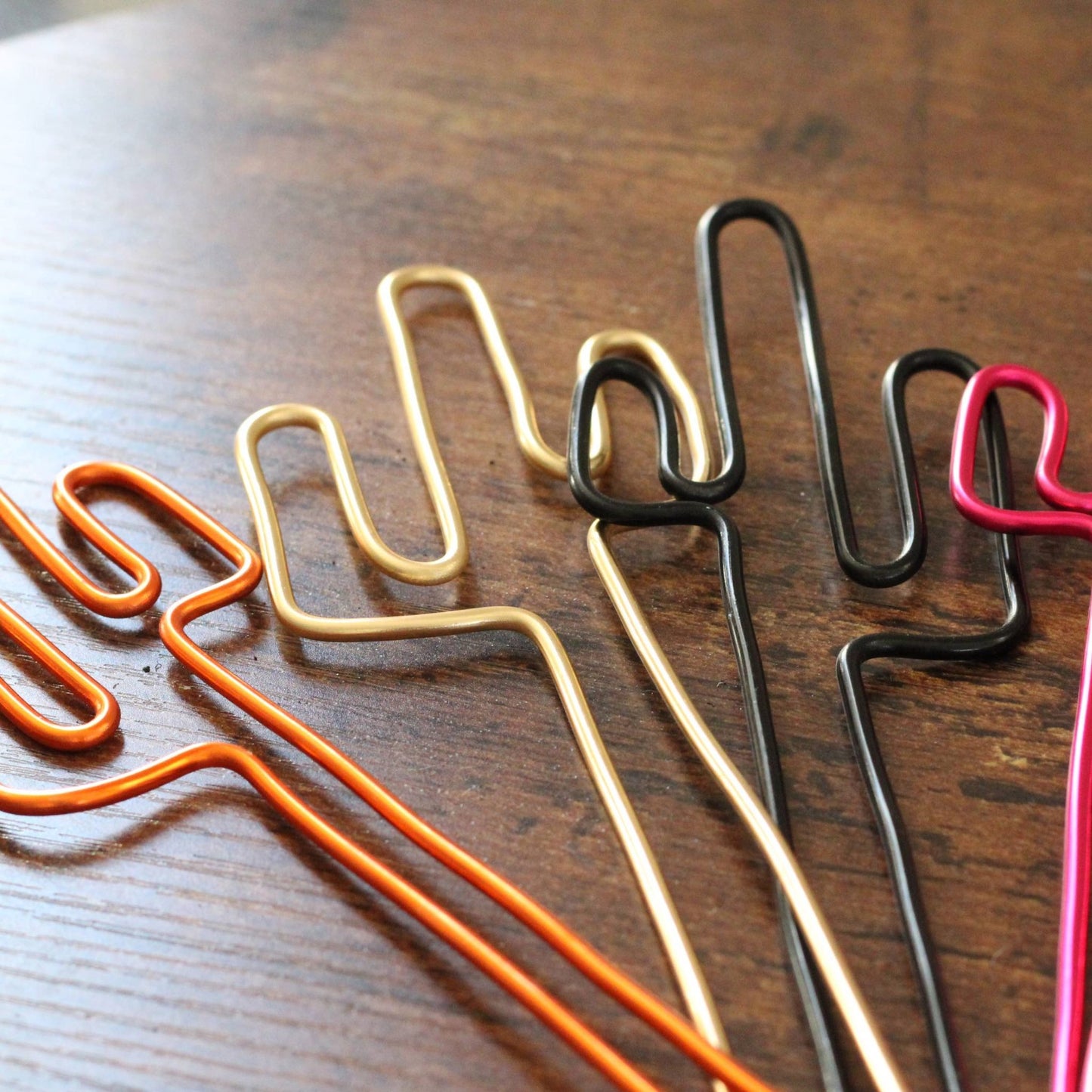 A variety of handcrafted cactus-shaped plant stakes in dark copper, gold, black, and bright pink aluminium; designed as decorative plant supports or unique gift card holders for sustainable gifting.