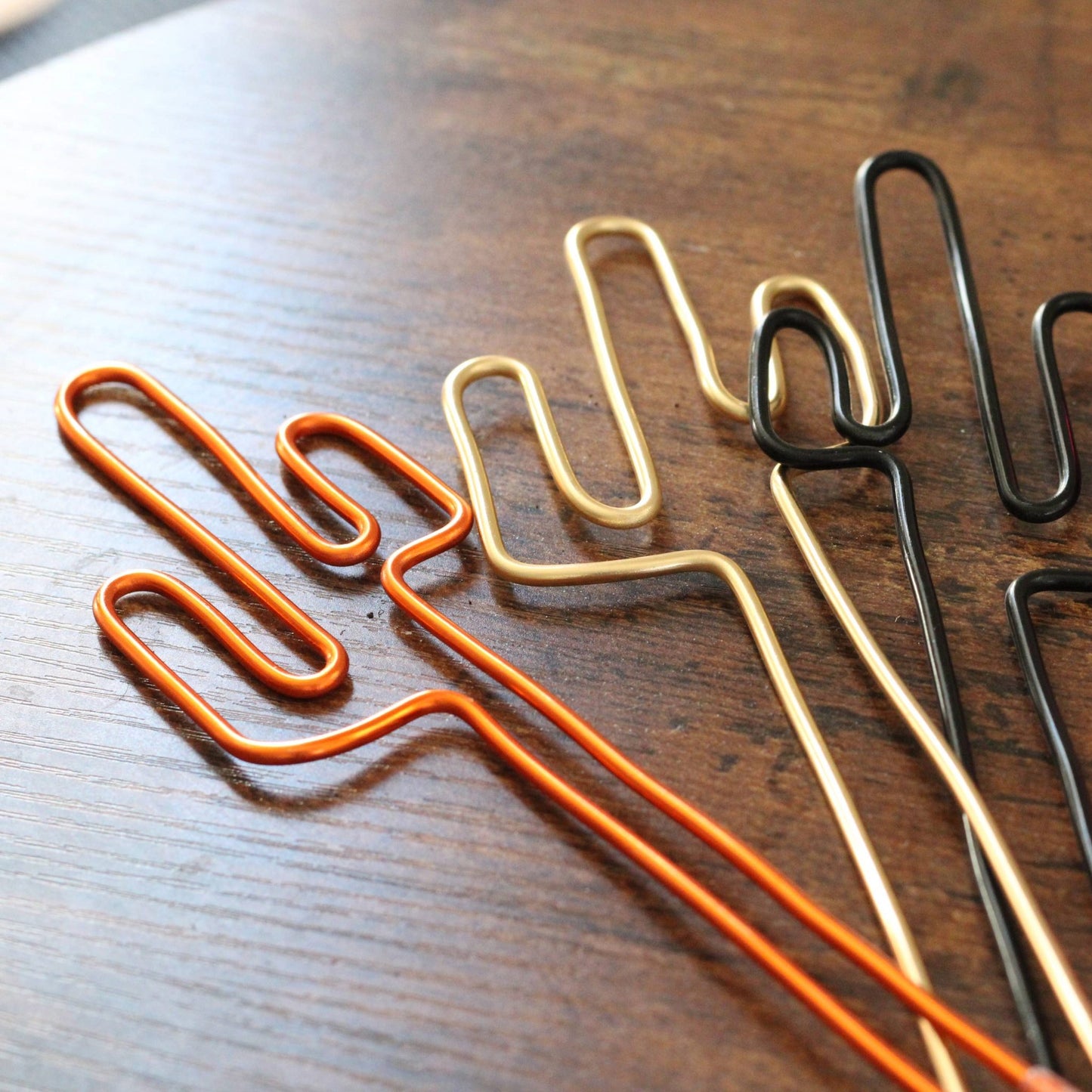 A variety of handcrafted cactus-shaped plant stakes in dark copper, gold, black, and bright pink aluminium; designed as decorative plant supports or unique gift card holders for sustainable gifting.