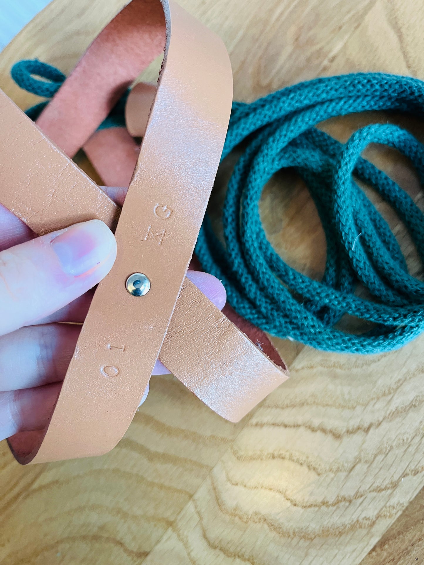 Hand holding a beige leather strap with '01 KG' branding on a wooden surface with green rope.